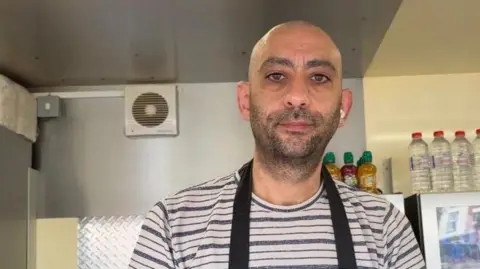 A man in a black and white striped T-shirt and a navy apron stands in a food truck. Behind him are lots of bottles of water and a transparent fridge with soft drinks in.