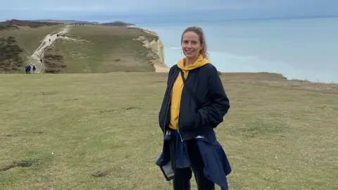 A blonde woman with a dark blue coat and a yellow hoodie, standing by a cliff.