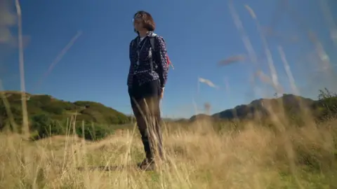 Botanist Heather Garrett stood in a field of tall yellow grass. She wears dark pants, a blue patterned long-sleeved top and a short dark bob and glasses.