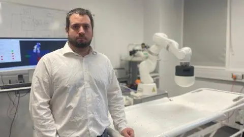 Bruno Scaglioni  stands in a laboratory wearing a white, open neck shirt.
Over his left shoulder is a computer screen and to his right is a robotics machine. 