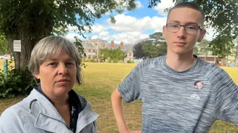 A woman in a striped anorak standing next to a teenager in a grey t-shirt and glasses with his arms on his hips with an old school building across the field behind them
