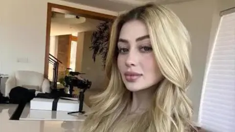 A selfie of a young woman with long blonde hair. She sits in a room with glossy stone surfaces and dark furniture.