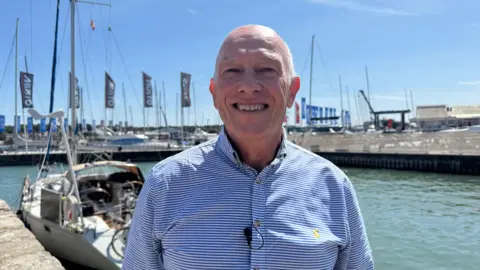 Image shows Jim smiling at the camera. He is wearing a blue and white striped shirt. Behind him are a number of flags and boats. He is stood on the edge of the harbour and at the bottom of the photo, you can see the sea.