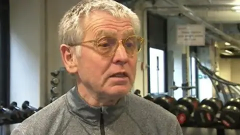 The shot of Peter Haynes is of his head and shoulders, he is wearing a grey training top and standing in a gym with free weights behind him 