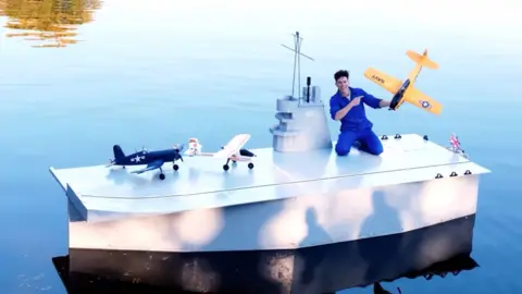 A man in a blue jumpsuit kneels on a white-coloured foam aircraft carrier on water while he holds a yellow remote-controlled plane in his left hand. Two other planes are on the deck of the carrier