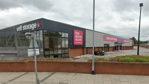 The long warehouse is lined with grey metal at the top, and red-brick at the bottom. Pink, purple and white signage erected on the side of the building reads: "The store room self storage." The building is set off the road behind a pavement, brick wall and line of hedges.