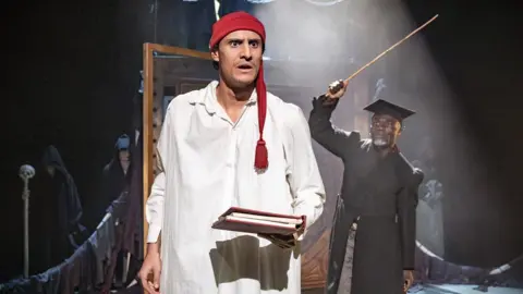 Craig Fuller A male actor is wearing Victorian nightgown performing as a character on stage with another male actor in a Victorian teacher's costume and a cane in his hand standing by a chalk board.