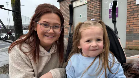 Demi Whelan and her daughter Tilly. Demi has long brown hair, wearing glasses and a cream fleece. Tilly has long blonde hair in pigtails and light blue jumper sitting in a pram. Demi is crouched down next to her.