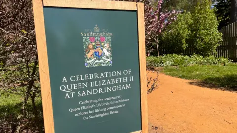 Shaun Whitmore/BBC An information board is on the left of the picture. It is beside a path. There are trees and shrubs in the background. The board is green and has a wooden frame. It reads: "A celebration of Queen Elizabeth II at Sandringham." 