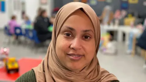 A woman in a brown headscarf and green top smiles at the camera.
