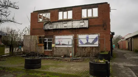The exterior of an derelict, red brick, two-storey building. It has boarded up windows on the ground floor and is surrounded by wire fencing.