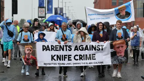 A group of people holding up an anti-knife crime banner walk in the rain outside Ibrox
