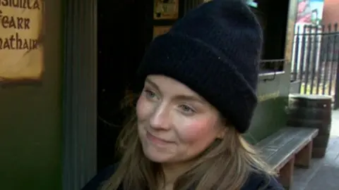 Claire Kieran has long brown hair and is wearing a black hat and black coat standing outside a Belfast pub with dark green walls.