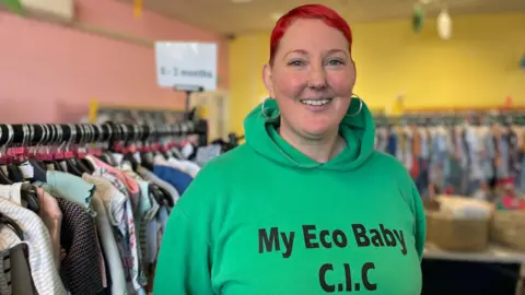 BBC/Gemma Dillon A woman with short red hair wearing a green hoody with the words "My Eco Baby CIC" stands in front of rails of clothes.