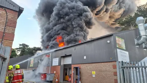 Hertfordshire Fire and Rescue Service Large plumes of dark smoke billowing out of a brick industrial building. There are some fire crews in the foreground and jets of water are aimed at the building.