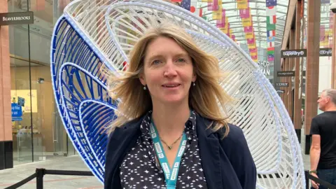BBC A blonde woman stands in a shopping mall with a modern art sculpt behind her. She wears a blue dress and cardigan.