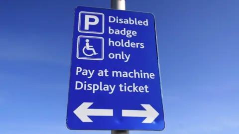 The image shows a blue sign identifying spaces for disabled badge holders and for pay at machine instructions in a car park. The sign says disabled badge holders only, pay at machine, display ticket. A blue sky is in the background.