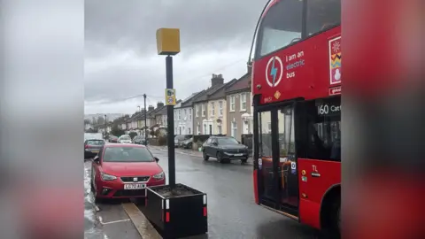 Fake yellow speed camera in a planter next to red double decker 160 bus in the foreground. Parked cars and houses can be seen in the background