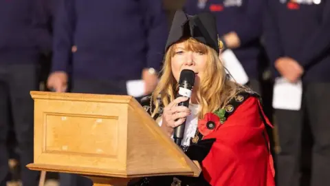 David Calvert The mayor of Aldeburgh TJ Halworth-Culf - a woman with long ginger/blonde hair dressed in a red mayoral gown and wearing a black hat. She is holding a mic. 