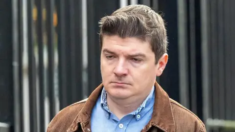 Michael McMonagle walking into court wearing a blue shirt and brown jacket. His hair is short and brown. The courthouse railings can be seen in the background