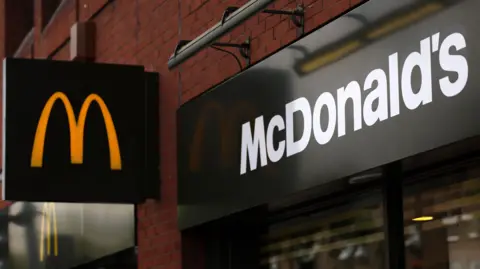 Shutterstock A exterior shot of a McDonald's restaurant with a black sign displaying a yellow "M" on it, with the full "McDonald's" title written above the entrance. 