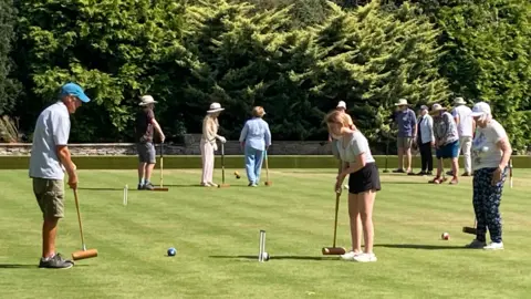 A group of players which consist of men, women and a teenager, are playing croquet on grass. The sun is shining and there are trees in the background. The players are holding mallets and are hitting three inch balls towards hoops pegged into the ground. Most are wearing hats, t-shirts, shorts and leggings with trainers. 