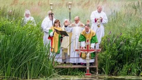 A group of people dipping a crucifix into the river in Tewkesbury. They are dressed in long white clergy robes and there are tall green reeds on the river bank