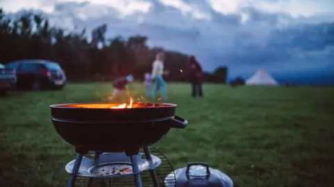 Getty Images A round, black charcoal kettle barbecue grill with a lit, glowing fire inside sits in the foreground of a grassy field. The grill is mounted on a metal tripod stand with a small tray underneath, and its lid rests on the grass nearby. In the soft, blurred background, several people stand near a tent under a dramatic, cloudy twilight sky, with a car parked on the far left.