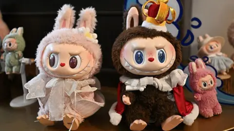 Labubu dolls of various sizes on a display shelf. At the centre of the image are a pastel pink and brown dolls, both with wide grins on their faces. The brown doll is wearing a crown and red 6cape 