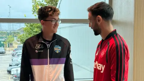 Family handout Zac face-to-face with Manchester United club captain Bruno Fernandes. They are standing by a window and smiling at each other. Fernandes is wearing a red sports top with black stripes on the arms, while Zac has a black, purple and blue sip up top on,