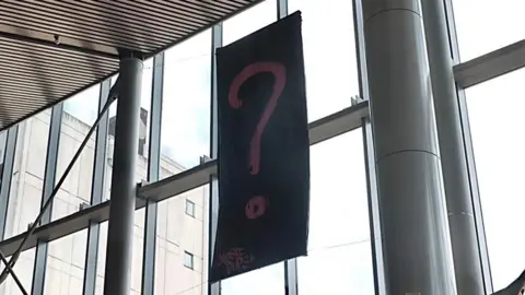 A large black banner with a pink question mark hangs in front of large windoes. Around it are metal support beams. 