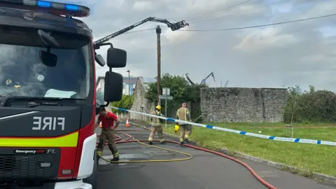 A fire engine is parked at the side of a residential street. A firefighter can be seen dealing with a hose at the side of the vehicle while two others step the other side of police tape onto a grass verge, heading towards the scene of the fire - a derelict hospital. There are hoses all over the floor and the sky is filled with smoke.