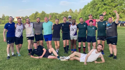 Rugby players pose for a group photo. Two are lying on their sides on the grass whilst they all cheer.