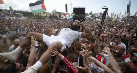 Getty Images Bobby Vylan, lead singer of Bob Vylan, is held aloft by fans as he lies out. at the front of the band's show at Glastonbury Festival 2025, on Saturday. He has a microphone to his mouth and is wearing white T-shirt and shorts with white socks and hi-top sneakers.