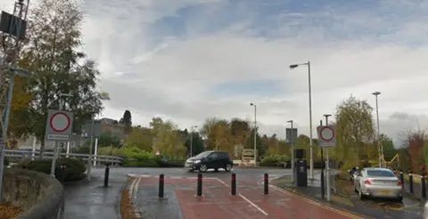 A roundabout with the road to the south of the photo pedestrianised with bollards. Parked cars to the right and a car is travelling through the roundabout