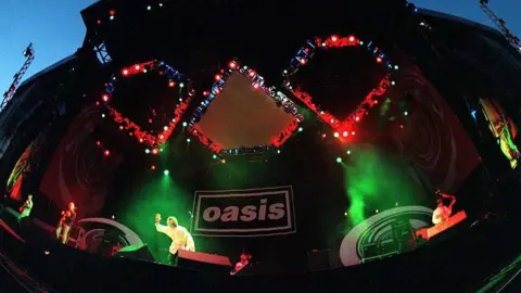 Getty Images Oasis on stage. Green stage lights illuminate them both, with a black backdrop with the Oasis logo on it.