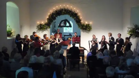 Maggie Faultless People playing instruments inside a building as a small crowd of people watch. It appears to be evening time and there are lighted candles above greenery which has been used to decorate the top of an arched doorway.