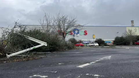 A rundown car park with the Toys R Us store visible behind bushes and trees. Structures which used to house trolleys can be seen toppled over on some of the bushes. Some cars are parked in the area.