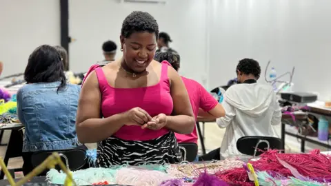 For the door of courtesy Danyi Mcletchie Danii McLertchie on a table filled with feathers and sequins. She looks down her hand while sorting through materials. 