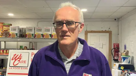 Fintan McGrath is standing in a foodbank with groceries behind him. He wears glasses and is wearing a shirt under a purple hoodie.