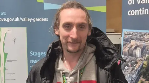 Nash Travelman A man with long hair and a goatee wearing a hoodie and a jacket. He is looking at the camera and smiling.