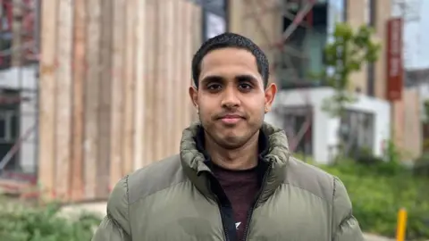Sachin Bhardwaj is wearing an olive-coloured padded jacket and a maroon top and is standing in front of The Gorge co-living block in Exeter