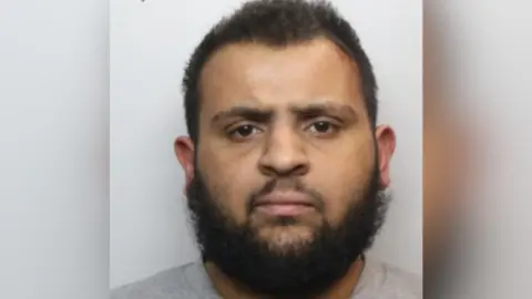 GMP A man in a mugshot with black hair and a black beard.
