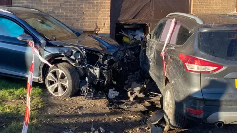 A car sitting parallel to a house. The wall of the property is cracked and has caved in. The front end of the car is crumpled and completely destroyed. The car's airbag has gone off and can be seen through one of the front windows.