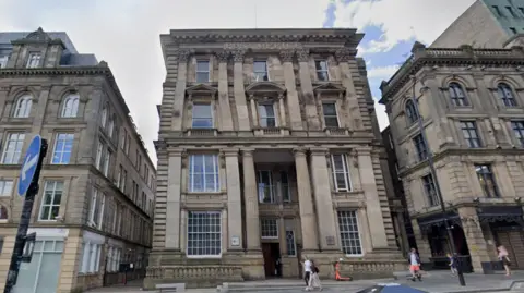 Large old white building with pillars and large windows. On the top of the multi-story brick building it has "Post Office" written in large lettering.