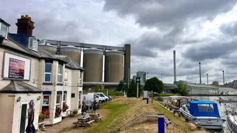 Martin Giles/BBC Sugar storage silos at Cantley sugar factory with a river in the foreground on a cloudy day. There are boats moored on the river and a pub on the left of the picture.