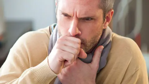A man in yellow jumper and grey scarf coughing