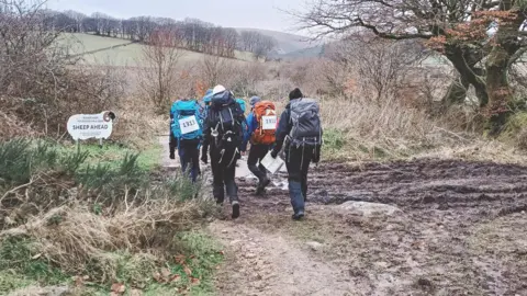 Four people walking over muddy ground in the wintry Devon countryside. They all have rucksacks on and are walking together.