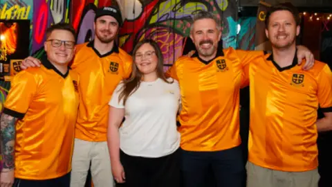 Old Gold Pack Kieran Newey is wearing one of the shirts and is standing with four other people. Three are wearing the shirt.