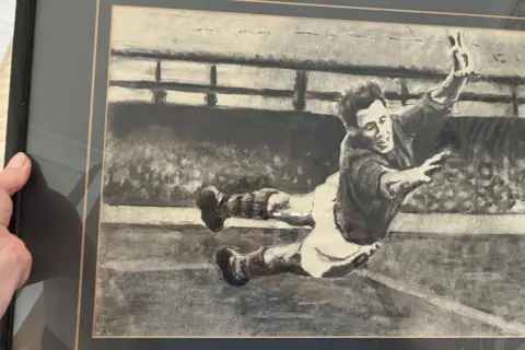 A framed charcoal drawing shows a young Roy Wood falling and reaching as he throws himself across goal to make a save in his Leeds United playing days in front of a stand full of people.